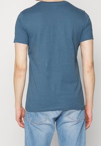 Persona che indossa una semplice camicia blu a maniche corte e jeans di denim blu chiaro, in piedi con la schiena rivolta verso l'osservatore su uno sfondo neutro.