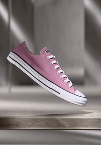 Pink canvas low-top sneaker with white rubber sole and toe cap. Features classic lace-up design and black stripe along the sole.