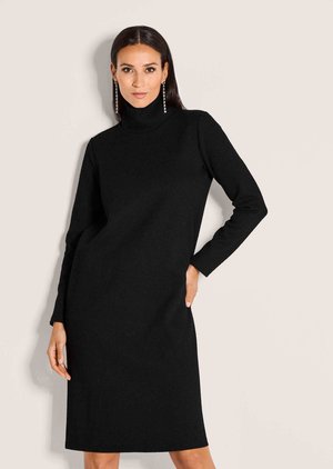 Frau mit langen dunklen Haaren, die ein schwarzes, langärmeliges, knielanges Kleid mit Rollkragen und lange hängende Ohrringe trägt, posiert vor einem schlichten Hintergrund.
