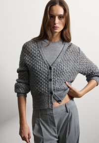 Grijze gebreide cardigan met een textuurpatroon, knoopsluiting aan de voorkant en geribde manchetten. Draagstijl over een grijze geribde top, gecombineerd met grijze broeken.
