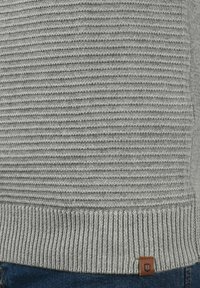 Grauer Strickpullover mit strukturierten horizontalen Streifen, geripptem Saum und einem kleinen braunen Lederlogo-Patch am unteren Ende.