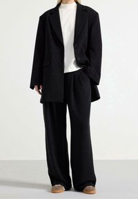 Blazer nero oversize con due bottoni, indossato sopra una maglietta bianca. Pantaloni neri a gamba larga e scarpe grigie slip-on completano il look.