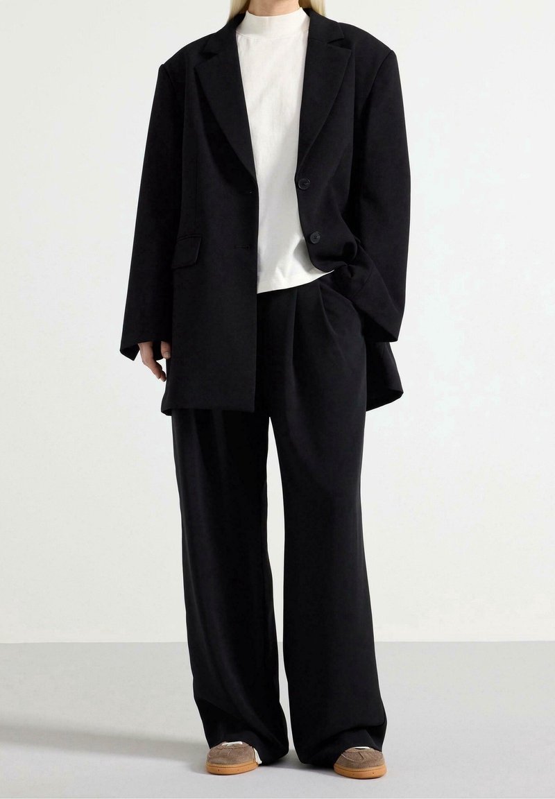 Blazer nero oversize con due bottoni, indossato sopra una maglietta bianca. Pantaloni neri a gamba larga e scarpe grigie slip-on completano il look.