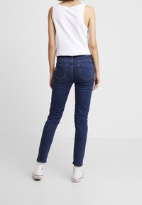 Jeans ajustados de color azul oscuro con una textura suave y bolsillos traseros, combinados con una camiseta sin mangas blanca y zapatillas blancas.