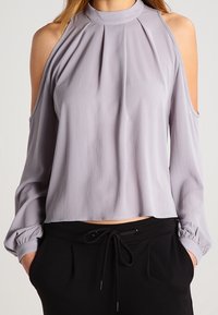 Blouse lavande claire à épaules dénudées, col plissé, manches longues et poignets élastiques. Associée à un pantalon noir à cordon de serrage.