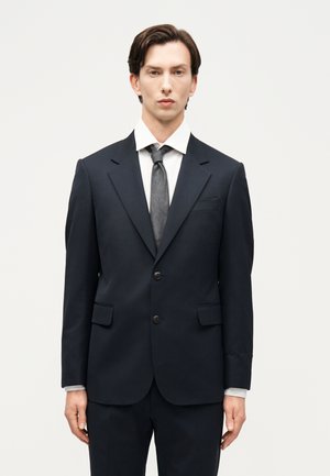 Άνδρας με κοντά σκούρα μαλλιά φοράει μαύρο κοστούμι, λευκό πουκάμισο και γκρι γραβάτα, στέκεται μπροστά σε λευκό απλό φόντο.