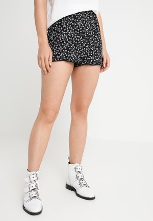 Persoon draagt zwarte korte broek met witte bloemprint, witte top en witte enkellaarzen met drie zilveren gespen aan elke laars.