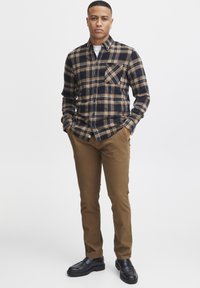 Camisa de franela a cuadros en azul marino y beige, con cuello abotonado y un bolsillo en el pecho. Combinada con pantalones beige y zapatos negros.
