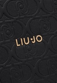 Fekete, textúráj�ú pénztárca, melyen dombornyomott minta található, és az elején arany színű "LIU·JO" logó díszíti. Simább anyag, amely finom fényt ad.