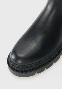 Primo piano della punta di un stivale in pelle nera con superficie texturizzata e suola in gomma spessa e resistente su sfondo chiaro.