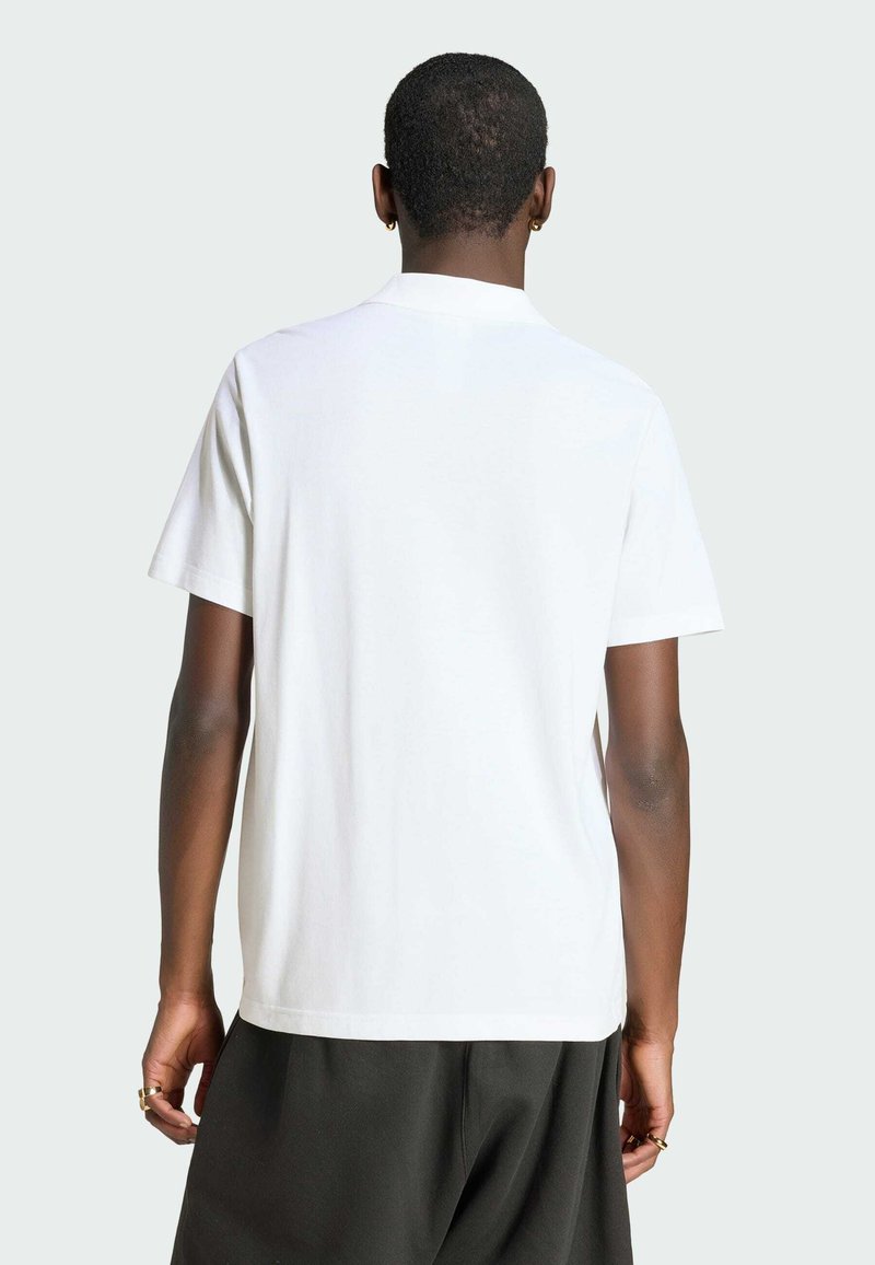 Person mit kurzen Haaren, die ein schlichtes weißes Kurzarmshirt und dunkle Hosen trägt, von hinten vor hellem Hintergrund gezeigt.