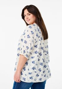 Blusa bianca con stampa floreale blu, tessuto morbido, scollatura rotonda e maniche corte arricciate. Abbinata a jeans blu.