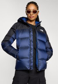 Marineblauwe en zwarte gewatteerde jas met capuchon, voorzien van een quilted ontwerp, een ritssluiting aan de voorkant en zijzakken. Het model draagt een zonnebril en een zwarte top.