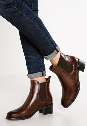 Bottes courtes en cuir marron avec panneaux latéraux élastiques, petit talon bloc, et texture lisse, associées à un jean foncé retroussé.