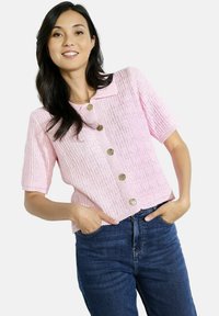 Gaiši rozā īsām piedurknēm krekls ar pogām, teksturētu dizainu un noapaļotu jostu, ar zeltītiem pogām un īsu piegriezumu, kombinēts ar ziliem džinsiem.