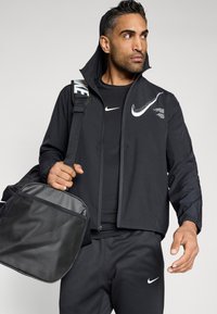 Μαύρο αδιάβροχο Nike με φερμουάρ, διακοσμημένο με λογότυπο, συνδυασμένο με μαύρο μπλουζάκι και παντελόνι, κρατώντας μια μαύρη τσάντα duffel με λεπτομέρεια από δίχτυ.