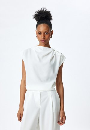Top bianco senza maniche con design drappeggiato e tre bottoni decorativi sulla spalla, abbinato a pantaloni bianchi coordinati in tessuto texturizzato.
