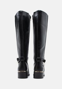 Mustat polven yli ulottuvat saappaat, joissa on teksturoitu pinta, sileät nahkaakoset, sivuvetoketjut ja pienet kultaiset yksityiskohdat. Tukevat kanta-muotoilut.