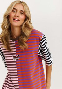 Tęczowa, pasiaste, oversize'owa koszulka wykonana z miękkiego materiału. Posiada naszywkę w kształcie motyla na różowym, fioletowym, czerwonym i czarnym pasku.