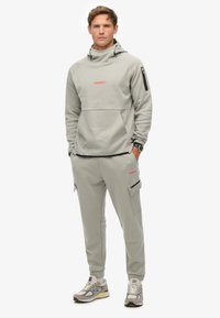 Sudadera gris claro con cuello alto y bolsillo frontal, combinada con pantalones ajustados a juego que tienen bolsillos con cremallera y detalles en negro.