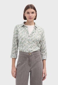 Camisa blanca de botones con patrones de plantas verdes, de manga larga y con cuello clásico, combinada con pantalones grises de cintura alta con bolsillos.