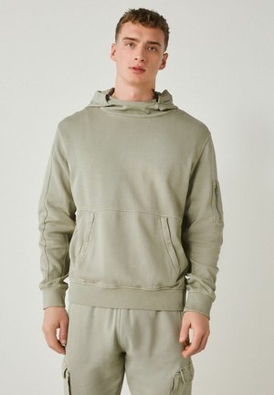 Junger Mann trägt einen hellgrünen Kapuzenpullover und passende Cargohosen und steht vor einem schlichten hellen Hintergrund.