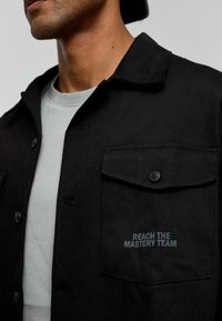 Zwarte denimjas met een kraag, knoopsluiting en twee borstzakken. Bevat geborduurde tekst "BEREIK HET VAKTEAM" op de zak.