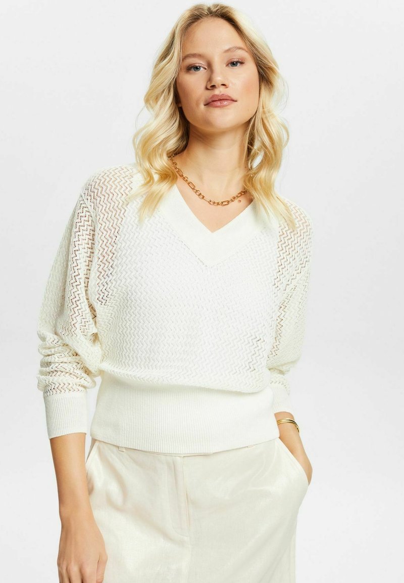Femme blonde portant un pull en tricot blanc avec col en V, un pantalon crème taille haute, un collier en chaîne dorée, et un bracelet doré avec une main dans la poche.