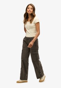 Beige Kurzarmtop mit Knopfdetails. Olivgrüne Cargo-Hosen mit mehreren Taschen und elastischen Bündchen. Hellbraune Sneakers.