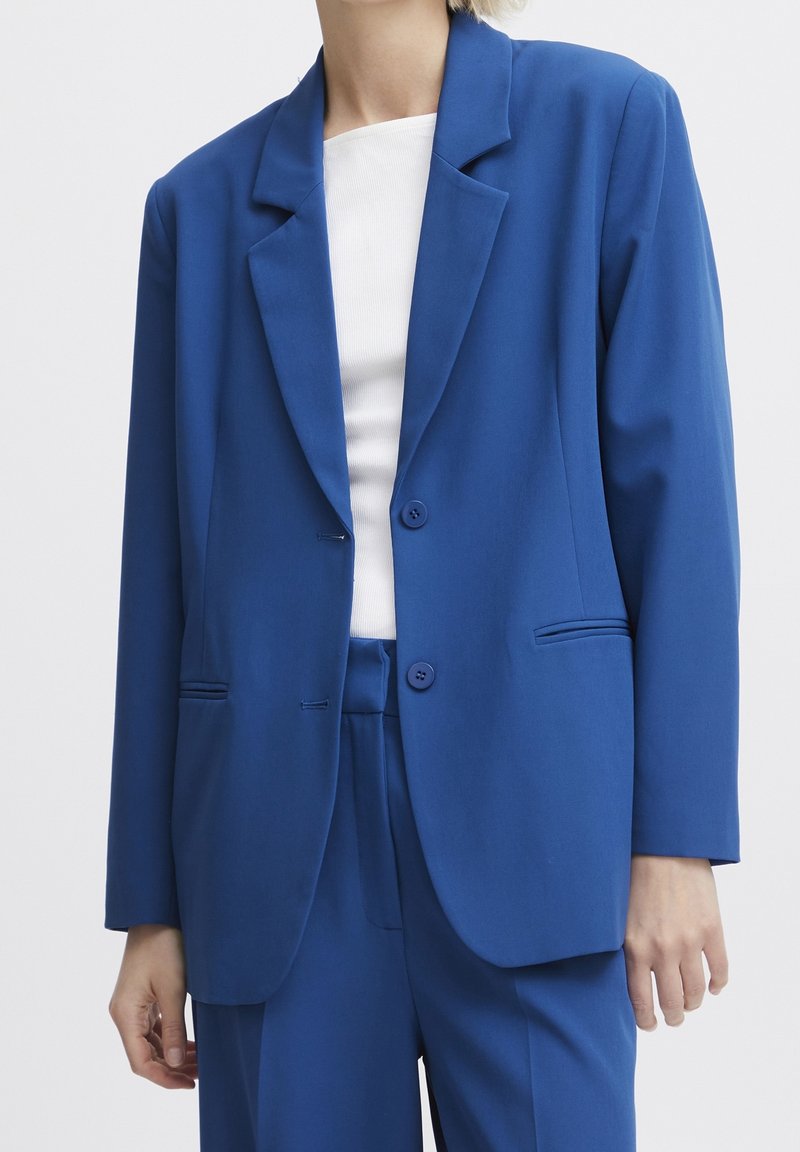 Personne portant une veste de costume bleu roi et un pantalon assorti avec un haut blanc dessous, debout devant un fond blanc uni.