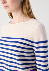 Femme portant un pull crème à rayures horizontales bleues et des boucles d'oreilles argentées texturées, vue des épaules au menton dans un éclairage doux.
