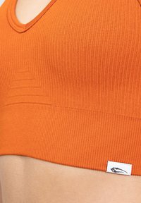 Sujetador deportivo de color óxido, con canalé y escote en V, patrón triangular texturizado en la parte delantera y una pequeña etiqueta blanca de la marca en el dobladillo.