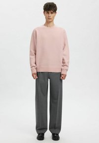 Lichtroze gebreide trui met een ronde hals, gecombineerd met wijde grijze broek en zwarte schoenen, op een neutrale achtergrond.