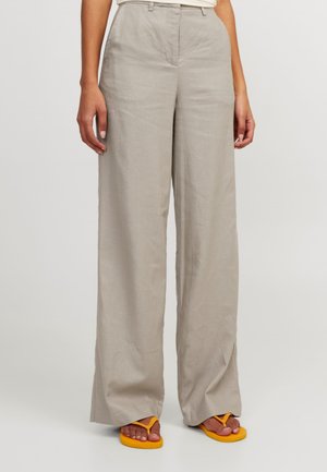 Beżowe spodnie z szerokimi nogawkami noszone przez osobę stojącą, zestawione z jasnopomarańczowymi klapkami na prostym jasnym tle.