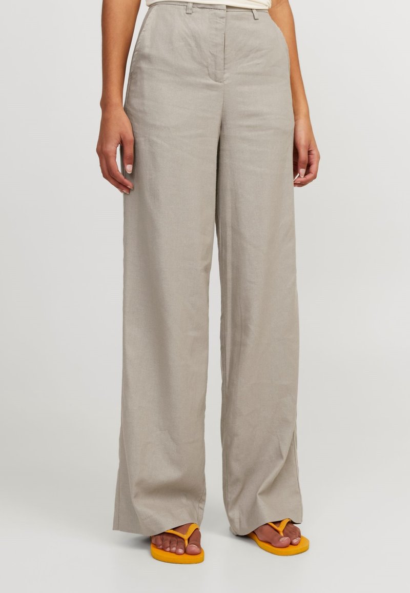 Pantalon beige à jambes larges porté par une personne debout, associé à des tongs orange vif sur un fond clair uni.