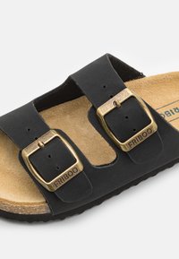 Svart sandal med to justerbare spenner, korkfotbed og strukturert gummisåle. Brun innersåle synlig med detaljer om merkevaren.