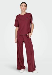 Bordeauxfarbener Sportanzug mit einem kurzärmeligen Oberteil und weißen Seitenstreifen sowie weiten Hosen. Hergestellt aus glattem, leichtem Stoff.