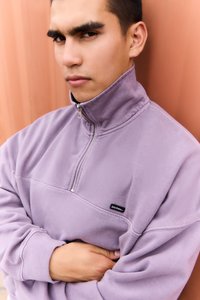 Světlý fialový sweatshirt na čtvrt zip s vysokým límcem, měkkou texturou a malou černou nášivkou na levé části hrudi, který má žebrované manžety.