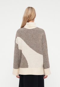 Sweter dzianinowy z wysokim kołnierzem, z pionowymi brązowymi i kremowymi paskami oraz dużym kremowym kształtem na plecach. Miękka tekstura, luźny krój.