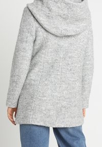 Cappotto in lana grigia e texturizzata con un ampio cappuccio, maniche lunghe e taglio dritto. Presenta dettagli delle cuciture lungo la schiena e il bordo inferiore.