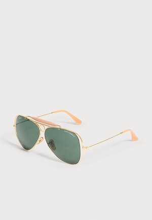 Γυαλιά aviator με χρυσό πλαίσιο και πράσινους φακούς, καφέ στηρίγματα μύτης και άκρες κροσσών σε χρώμα ροδάκινο, εμφανίζονται σε φωτεινό φόντο.
