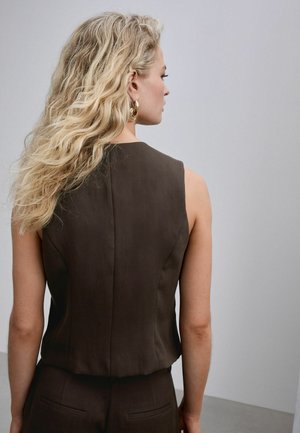 Woman with long blonde wavy hair wears sleeveless dark brown top and matching pants, facing sideways against a plain light background.