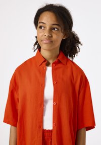Overmaatse rode linnen shirt met korte mouwen, voorzien van een knoopsluiting aan de voorkant en een ontspannen kraag, gedragen over een witte geribbelde tanktop.
