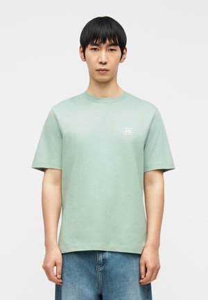 Mężczyzna ubrany w jasnozieloną koszulkę z krótkim rękawem z małym białym logo na lewej piersi oraz niebieskie dżinsy, stojący na tle białego, gładkiego tła.