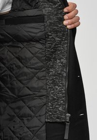 Černá podšívka bundy s prošívaným černým vnitřkem a texturovanou šedou látkou. Obsahuje zip s černými detaily a těsný střih.