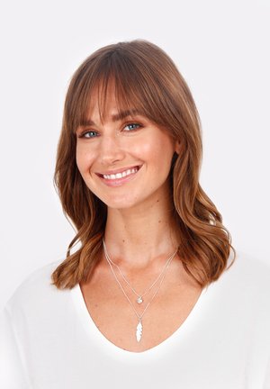 Kārtotas sudraba kaklarotas ar lapas piekariņu un mazāku apaļu kulonu, valkātas pāri baltam V-veida kakla izgriezumam, akcentējot gludas tekstūras un smalku dizainu.