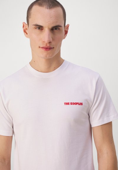 Camiseta de algodón blanca con cuello redondo, que presenta un logo bordado en rojo "THE KOOPLES" en la parte izquierda del pecho. Diseño minimalista.