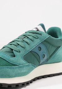 Baskets vertes mettant en avant un mélange de matériaux en daim et en mesh, avec des lacets texturés, un col rembourré et une semelle en caoutchouc de couleur claire.