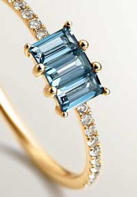 Anillo de oro con tres piedras azules de corte rectangular, adornado con pequeñas piedras transparentes a lo largo de la banda para un detalle adicional. Textura de superficie suave.