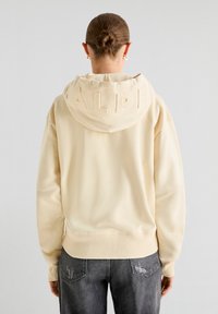 Crème-kleurige hoodie met een grote, gestructureerde capuchon en geribbelde manchetten, gecombineerd met donkergrijze versleten jeans. Glad materiaal met minimale details.